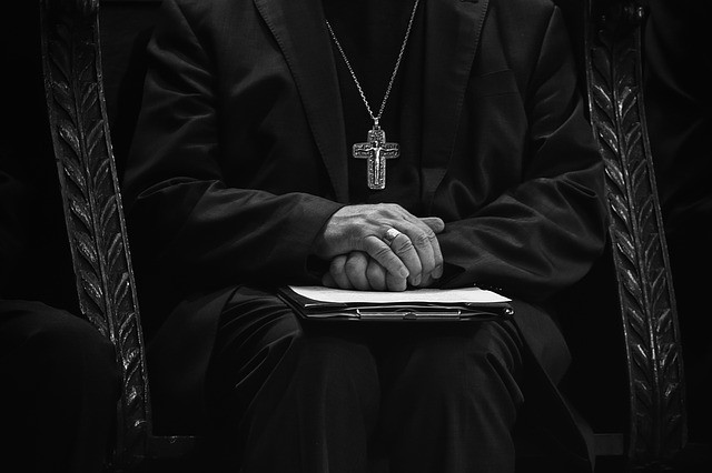 A Church official/cleric in black and white, face unseen, sitting, holding some documents